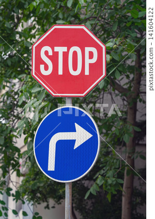 Red stop sign above a blue mandatory right turn sign against green foliage. Urban road signs representing traffic regulations, driving rules, road safety and direction Red stop sign above a blue mandatory right turn sign against green foliage. Urban road signs representing traffic regulations, driving rules, road safety and direction 141604122