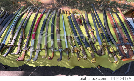Antique Swords and Sabers. Large collection of antique swords, sabers, decorative blades, scabbards on market table. Historic edged weapons feature ornate hilts, metal blades and aged patina Antique Swords and Sabers. Large collection of antique swords, sabers, decorative blades, scabbards on market table. Historic edged weapons feature ornate hilts, metal blades and aged patina 141604159