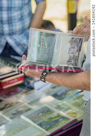 Woman Browsing Vintage Postcards in Collectors Album. Woman holds album containing vintage postcards, browsing collectibles at market stall. Nostalgia, personal memories, postcard collecting Woman Browsing Vintage Postcards in Collectors Album. Woman holds album containing vintage postcards, browsing collectibles at market stall. Nostalgia, personal memories, postcard collecting 141604192
