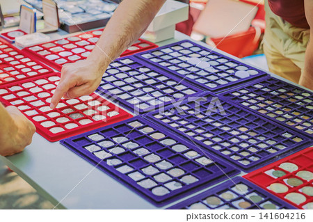 Collector points at coin specimens arranged in display trays during marketplace transaction. Concept of flea market, coin collecting, numismatics, investment, trade, history, valuation Collector points at coin specimens arranged in display trays during marketplace transaction. Concept of flea market, coin collecting, numismatics, investment, trade, history, valuation 141604216