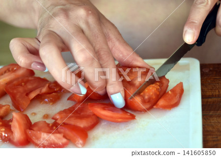 Close-up of a woman's hands cutting fresh tomatoes on a food-safe plastic cutting board while preparing a homemade meal in the kitchen. Close-up of a woman's hands cutting fresh tomatoes on a food-safe plastic cutting board while preparing a homemade meal in the kitchen. 141605850