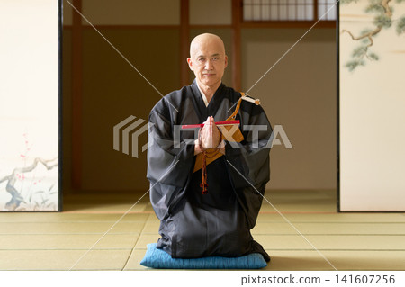 A monk sits in a kneeling position, hands clasped in prayer, holding prayer beads in his hand. A monk sits in a kneeling position, hands clasped in prayer, holding prayer beads in his hand. 141607256