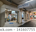 The station square walkway shows the ticket gates on the left and a shopping street extending to the right. 141607755
