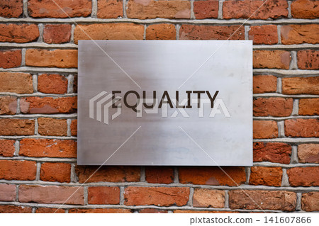 The sign with the word equality is placed on a metal plate attached to a brick wall. The setting appears to be in a city location during daylight The sign with the word equality is placed on a metal plate attached to a brick wall. The setting appears to be in a city location during daylight 141607866