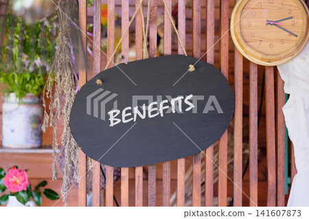 A sign hangs at a market stall showing the word benefits. The stall features wooden elements and has plants in containers nearby A sign hangs at a market stall showing the word benefits. The stall features wooden elements and has plants in containers nearby 141607873