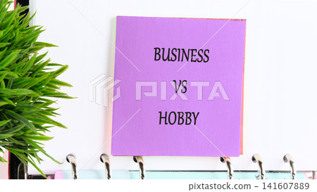 A purple sticky note shows the words business versus hobby on a desk. A small green plant is nearby. The scene suggests a conversation about choices related to work and leisure A purple sticky note shows the words business versus hobby on a desk. A small green plant is nearby. The scene suggests a conversation about choices related to work and leisure 141607889