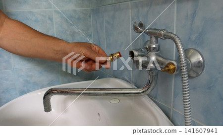 A technician is about to install a new valve body into a disassembled faucet. He holds a brass part with a red seal in his hand. The plumbing renovation is taking place in a bathroom with blue tiles. A technician is about to install a new valve body into a disassembled faucet. He holds a brass part with a red seal in his hand. The plumbing renovation is taking place in a bathroom with blue tiles. 141607941