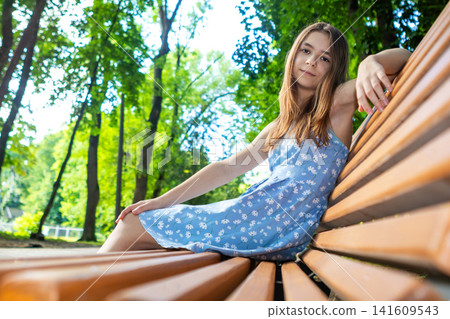 Teen girl relaxing on a wooden bench in a sunny green park wearing a blue summer dress Teen girl relaxing on a wooden bench in a sunny green park wearing a blue summer dress 141609543
