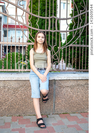 Teen girl sitting on stone ledge by decorative fence in casual summer outfit Teen girl sitting on stone ledge by decorative fence in casual summer outfit 141609544