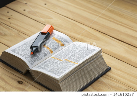 Open thick English dictionary book with orange highlighter marker pen resting on pages showing highlighted vocabulary words on rustic wooden table surface for learning and language education concept Open thick English dictionary book with orange highlighter marker pen resting on pages showing highlighted vocabulary words on rustic wooden table surface for learning and language education concept 141610257