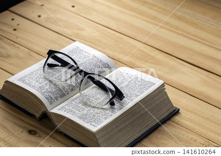 Close up of black framed reading eyeglasses resting on open vintage dictionary book with detailed text pages on rustic wooden plank table, symbolizing study, education and myopia concept Close up of black framed reading eyeglasses resting on open vintage dictionary book with detailed text pages on rustic wooden plank table, symbolizing study, education and myopia concept 141610264