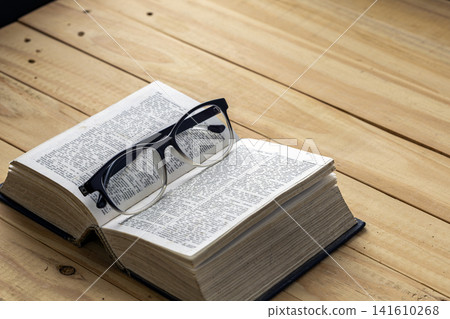 Black framed reading eyeglasses resting on open vintage dictionary book pages placed on rustic wooden plank table, concept of nearsightedness, education, learning, literature and knowledge study Black framed reading eyeglasses resting on open vintage dictionary book pages placed on rustic wooden plank table, concept of nearsightedness, education, learning, literature and knowledge study 141610268