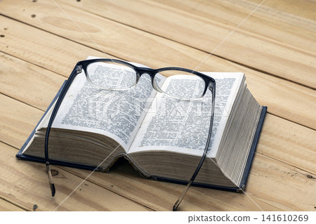 Open English dictionary book with black framed reading eyeglasses resting on top of pages placed on rustic wooden table surface, concept of education, learning, knowledge and nearsighted vision Open English dictionary book with black framed reading eyeglasses resting on top of pages placed on rustic wooden table surface, concept of education, learning, knowledge and nearsighted vision 141610269