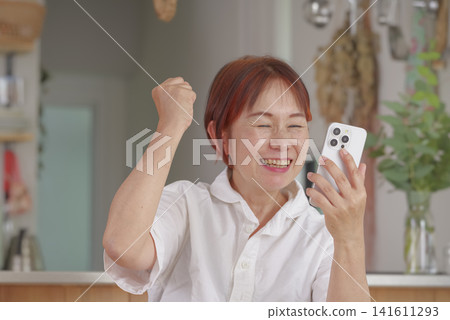 Middle-aged women making a triumphant gesture while looking at their smartphones. Middle-aged women making a triumphant gesture while looking at their smartphones. 141611293