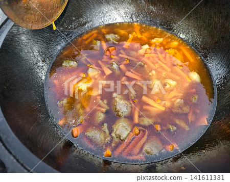 Cooking pilaf takes place in a kazan with meat and vegetables. A person pours ingredients into the pot while it warms over an outdoor flame Cooking pilaf takes place in a kazan with meat and vegetables. A person pours ingredients into the pot while it warms over an outdoor flame 141611381