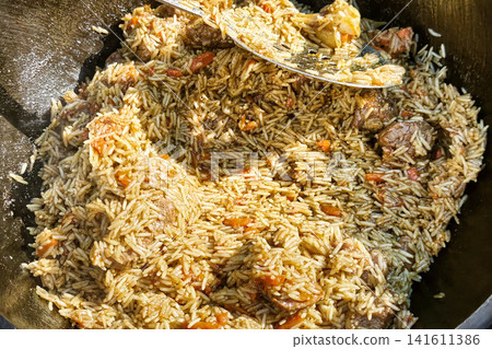People are preparing a large pot of rice with meat and vegetables in a kazan during a gathering in a sunny outdoor setting. The dish is being cooked carefully People are preparing a large pot of rice with meat and vegetables in a kazan during a gathering in a sunny outdoor setting. The dish is being cooked carefully 141611386