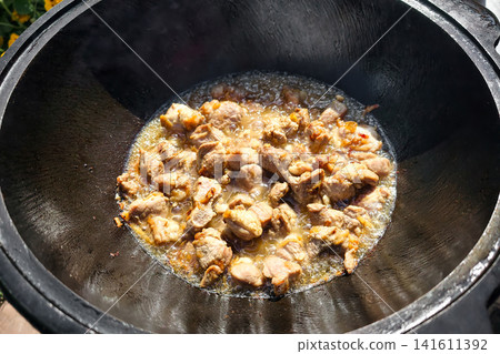 A kazan sits over a fire as meat cooks with spices. The setting is outside, showing the process of making pilaf. Steam rises from the dish A kazan sits over a fire as meat cooks with spices. The setting is outside, showing the process of making pilaf. Steam rises from the dish 141611392