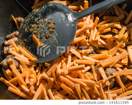 Slices of carrots mix with pieces of meat in a kazan. Cooking plov takes place at an outdoor kitchen with a fire. The process is focused and lively Slices of carrots mix with pieces of meat in a kazan. Cooking plov takes place at an outdoor kitchen with a fire. The process is focused and lively 141611395
