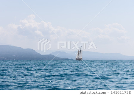 Sailboat on Lake Garda seen from the water on a summer day, blue waves stretch across the foreground while hazy mountain shores and large white clouds create an airy travel landscape with calm open Sailboat on Lake Garda seen from the water on a summer day, blue waves stretch across the foreground while hazy mountain shores and large white clouds create an airy travel landscape with calm open 141611738