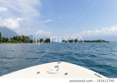 Summer boat trip on Lake Garda seen over the bow, rippling blue water opens toward green shores hazy mountains and a distant island for a peaceful vacation landscape with wide airy sky Summer boat trip on Lake Garda seen over the bow, rippling blue water opens toward green shores hazy mountains and a distant island for a peaceful vacation landscape with wide airy sky 141611740