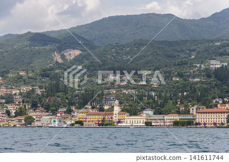 Gardone Riviera lakeshore beneath lush mountain slopes, elegant waterfront buildings cypress trees and hillside villas frame rippling Lake Garda in a scenic Lombardy resort view from the water 141611744