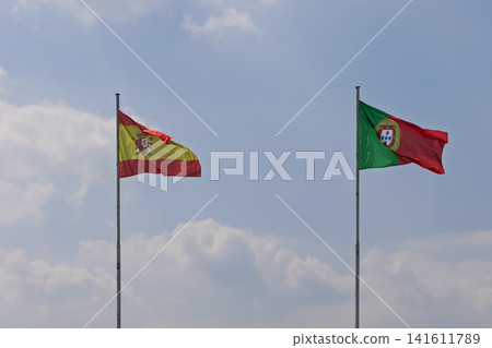 Flags of Spain and Portugal framing white clouds beneath a blue sky in daylight Flags of Spain and Portugal framing white clouds beneath a blue sky in daylight 141611789