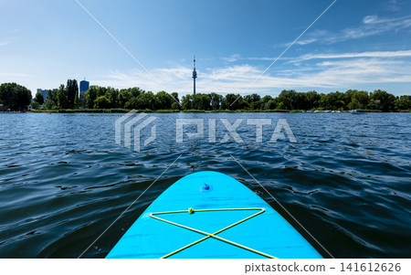 Turquoise paddleboard on water with modern city skyline, illustrating a relaxed summer water sport experience. Turquoise paddleboard on water with modern city skyline, illustrating a relaxed summer water sport experience. 141612626