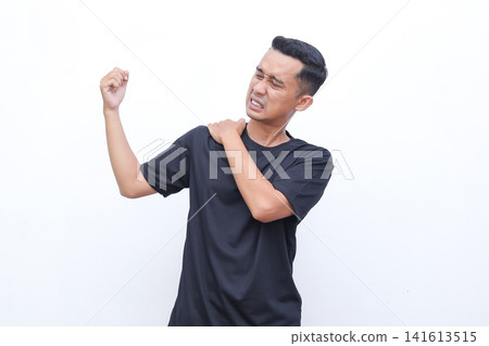 Man grips shoulder, showing pain. His face shows discomfort, eyes squeezed shut. He wears a plain black t-shirt. White background isolates the subject clearly. Ideal for health, fitness 141613515