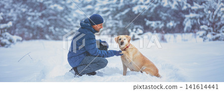 A human and dog are best friends. The man with the dog sitting in a snowy pine wood in winter. Trained Labrador retriever dog extends the paw to the man A human and dog are best friends. The man with the dog sitting in a snowy pine wood in winter. Trained Labrador retriever dog extends the paw to the man 141614441