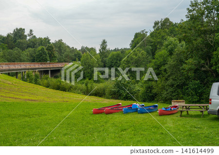 Colorful Canoes Resting on Green Riverbank Near Bridge in Peaceful Latvian Countryside Summer Colorful Canoes Resting on Green Riverbank Near Bridge in Peaceful Latvian Countryside Summer 141614499
