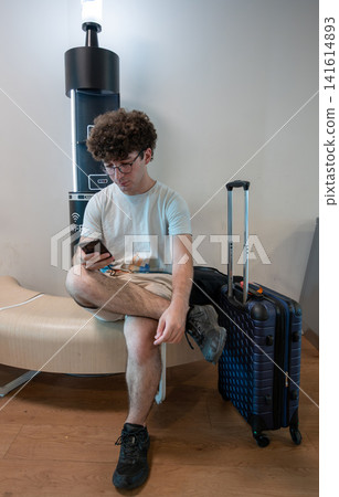 Young man waiting at the airport gate with luggage, connecting to wi fi and scrolling on his mobile phone while preparing for his journey 141614893