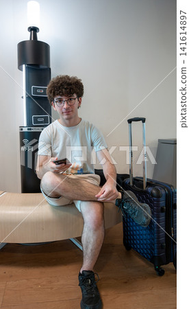 Young man with curly hair and glasses sitting by a charging station in an airport lounge, checking his smartphone while waiting with luggage Young man with curly hair and glasses sitting by a charging station in an airport lounge, checking his smartphone while waiting with luggage 141614897