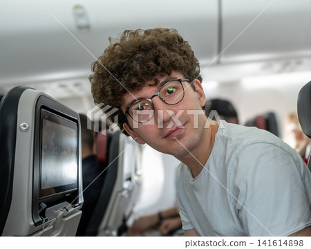 Young man with curly hair and glasses looking directly at the camera while traveling in the passenger cabin of an airplane, offering a friendly expression during his flight journey Young man with curly hair and glasses looking directly at the camera while traveling in the passenger cabin of an airplane, offering a friendly expression during his flight journey 141614898