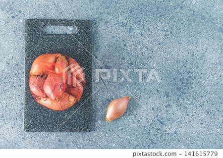 Fresh raw shallot on kitchen utensils background. Stone cutting board on dark grey table surface Fresh raw shallot on kitchen utensils background. Stone cutting board on dark grey table surface 141615779