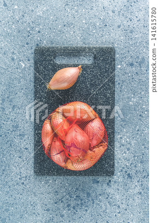 Fresh raw shallot on kitchen utensils background. Stone cutting board on dark grey table surface Fresh raw shallot on kitchen utensils background. Stone cutting board on dark grey table surface 141615780