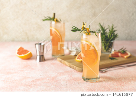 Two highball glasses of Paloma cocktails with tequila fresh grapefruit juice combined and rosemary. Surrounded of ingredients and bar tools. Light pink concrete surface. Two highball glasses of Paloma cocktails with tequila fresh grapefruit juice combined and rosemary. Surrounded of ingredients and bar tools. Light pink concrete surface. 141615877