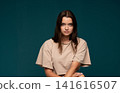 Young woman with serious expression looking at camera, posing for studio portrait with teal background 141616507