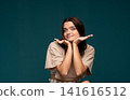 Young pretty woman with healthy skin smiling joyfully, posing with hands cupping face on a turquoise background 141616512