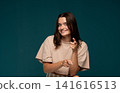 Young woman smiling, making a playful choosing gesture, pointing both index fingers forward against a solid teal background 141616513