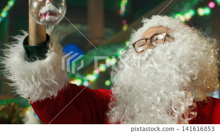 Professional Santa decorating his workshop with Christmas globe, preparing it for photographs with visitors. Elderly man in costume putting ornaments in room for holiday ambiance, camera A close up Professional Santa decorating his workshop with Christmas globe, preparing it for photographs with visitors. Elderly man in costume putting ornaments in room for holiday ambiance, camera A close up 141616853