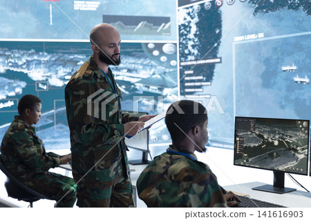 Military officer briefing personnel seated at command post workstation desks. Army squad team leader being provided mission information and intel data gathered by operators Military officer briefing personnel seated at command post workstation desks. Army squad team leader being provided mission information and intel data gathered by operators 141616903