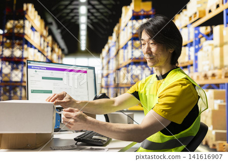 Worker with hi vis vest applying adhesive shipping labels with awb numbers on cardboard boxes, ensuring easy package tracking service on pc software. Manages labeling and packaging operations. 141616907