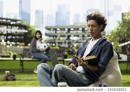Young asian man reading open book and taking notes on laptop, outdoor learning on university campus, writing down and studying alone on beanbag. Study time with stacked books. Young asian man reading open book and taking notes on laptop, outdoor learning on university campus, writing down and studying alone on beanbag. Study time with stacked books. 141616921