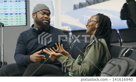 Black lovers talking about journey and waiting for flight departure, seated in airport lounge with runway view. Couple enjoying cute romantic moment, discussing leisure travel plan. Black lovers talking about journey and waiting for flight departure, seated in airport lounge with runway view. Couple enjoying cute romantic moment, discussing leisure travel plan. 141616940