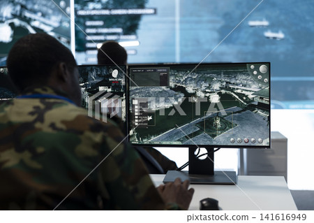 Man in military command post working on PC, looking at surveillance footage on monitor. Military operator watches intelligence feed at workstation desk, close up shot Man in military command post working on PC, looking at surveillance footage on monitor. Military operator watches intelligence feed at workstation desk, close up shot 141616949