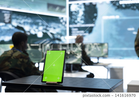 Close up of green screen tablet on military command post table in front of soldier working. Mockup device next to army personnel at workstations across operations room Close up of green screen tablet on military command post table in front of soldier working. Mockup device next to army personnel at workstations across operations room 141616970
