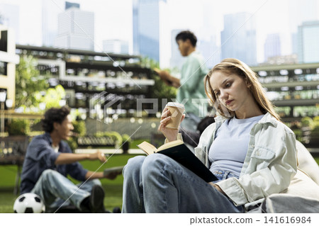 Woman student reading open book while holding coffee on beanbag in modern campus courtyard with classmates and study activity in background. Independent self study. Woman student reading open book while holding coffee on beanbag in modern campus courtyard with classmates and study activity in background. Independent self study. 141616984