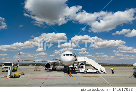 Commercial passenger airplane parked on airport tarmac. Ground crew servicing aircraft plane with boarding stairs and vehicles under blue sky with clouds. Commercial passenger airplane parked on airport tarmac. Ground crew servicing aircraft plane with boarding stairs and vehicles under blue sky with clouds. 141617058