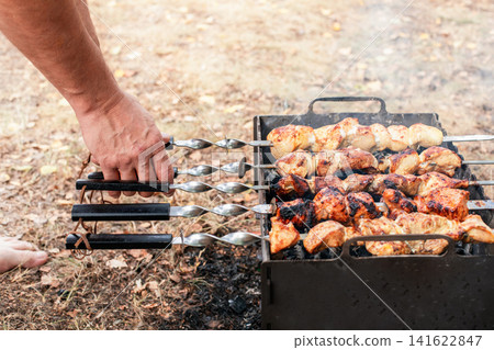 Close up of a person turning pork and chicken shish kebab skewers grilling over hot charcoal on an outdoor BBQ mangal. Close up of a person turning pork and chicken shish kebab skewers grilling over hot charcoal on an outdoor BBQ mangal. 141622847