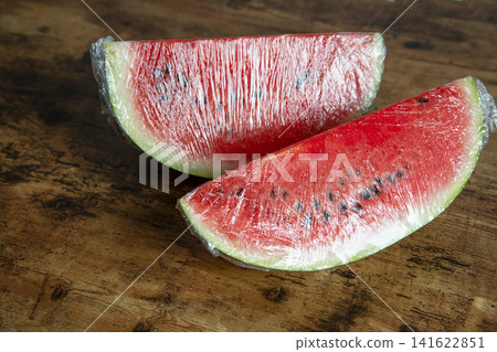 Two juicy red watermelon wedges covered with transparent stretch wrap on a rustic dark wooden table. Two juicy red watermelon wedges covered with transparent stretch wrap on a rustic dark wooden table. 141622851